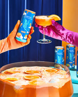 Two hands in colorful gloves holding a can and a cocktail glass with orange slices, surrounded by more cans and a punch bowl filled with orange-themed drinks.